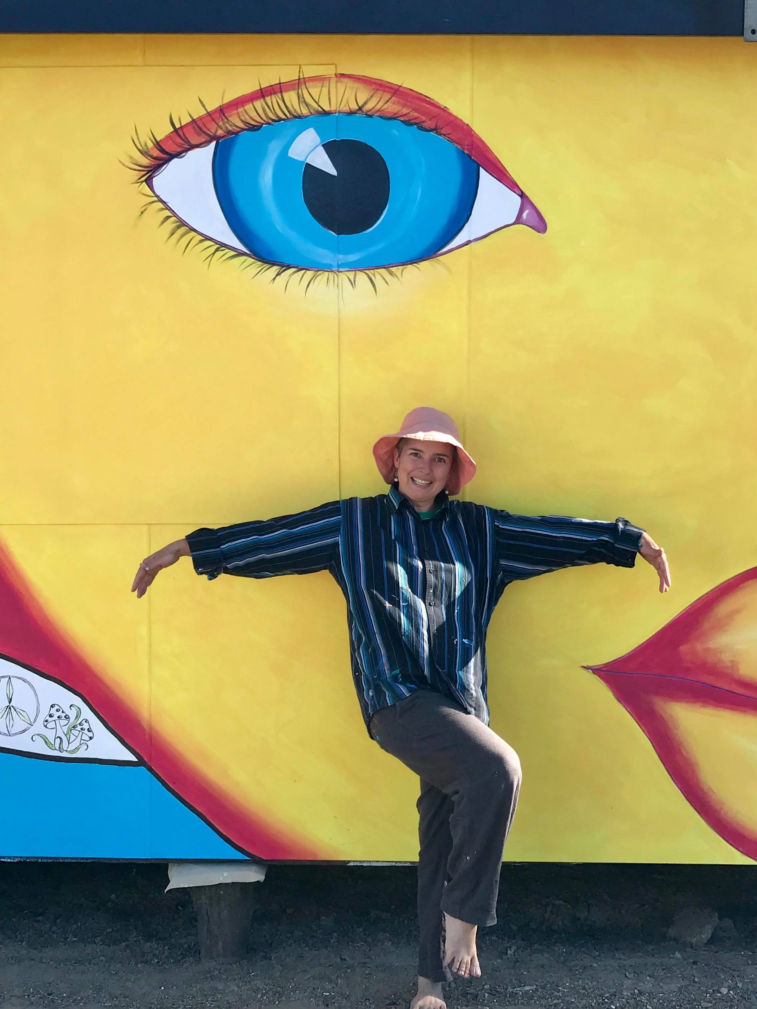A woman in a pink hat and striped shirt posing with arms outstretched in front of a colorful mural of a large blue eye with red and yellow accents.