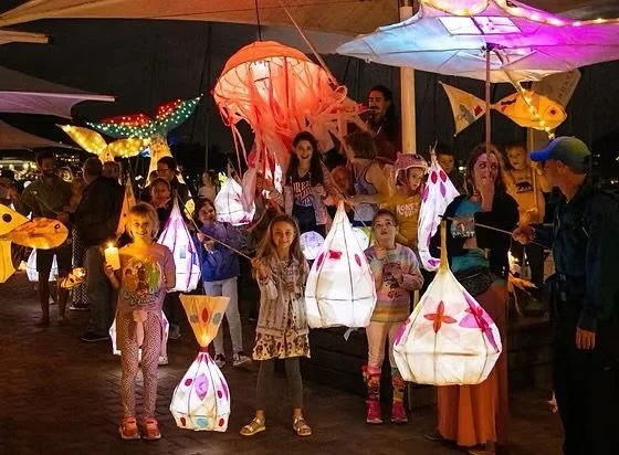 Children and adults at a lantern festival, holding and surrounded by colorful illuminated paper lanterns and decorative lights.