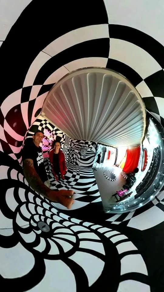 A fisheye view of a room with black and white spiral and checkerboard patterns on the walls and floors, with two people standing near the center, and red and black decor in the background.