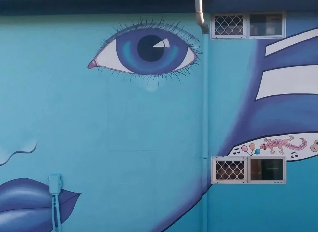 A mural of a large blue eye painted on a blue wall with two small windows and a door behind bars, and a design below the eye including balloons, a lizard, music notes, and other shapes.