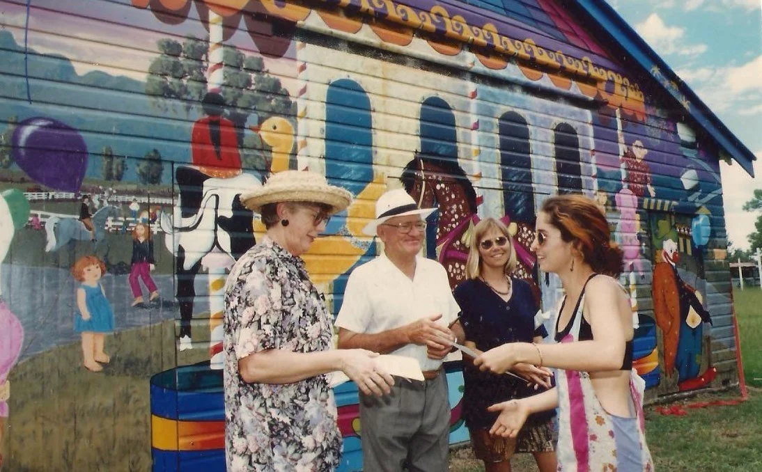 Four people standing outdoors in front of a colorful mural, engaging in conversation. One woman is wearing a large sunhat and floral blouse, another man has a hat and glasses, a woman in sunglasses and dark clothing, and a young woman with reddish hair in a colorful dress.