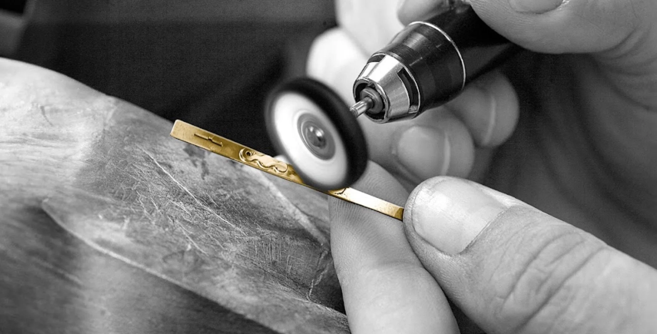 A person using a rotary tool to engrave a gold-colored metal piece, with focus on the tool and metal surface.