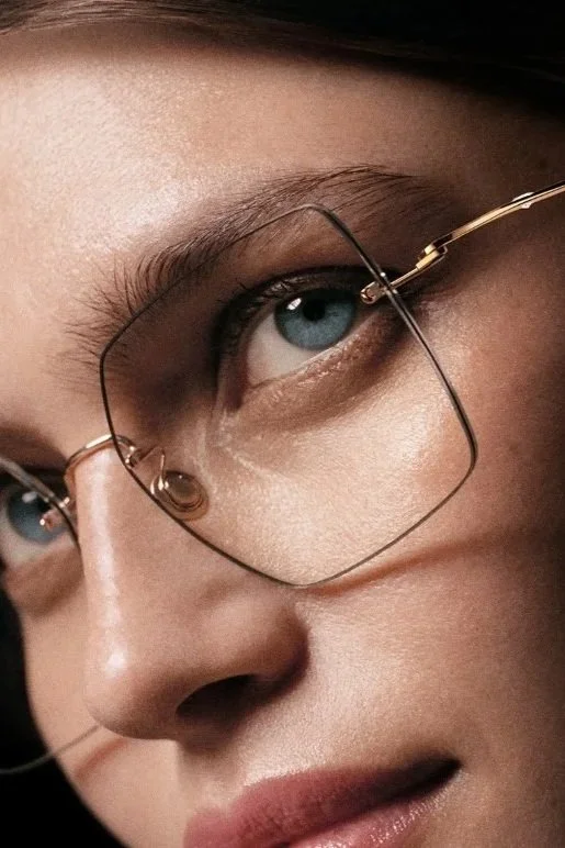 Close-up of a woman wearing large, square, gold-rimmed eyeglasses and staring directly ahead.