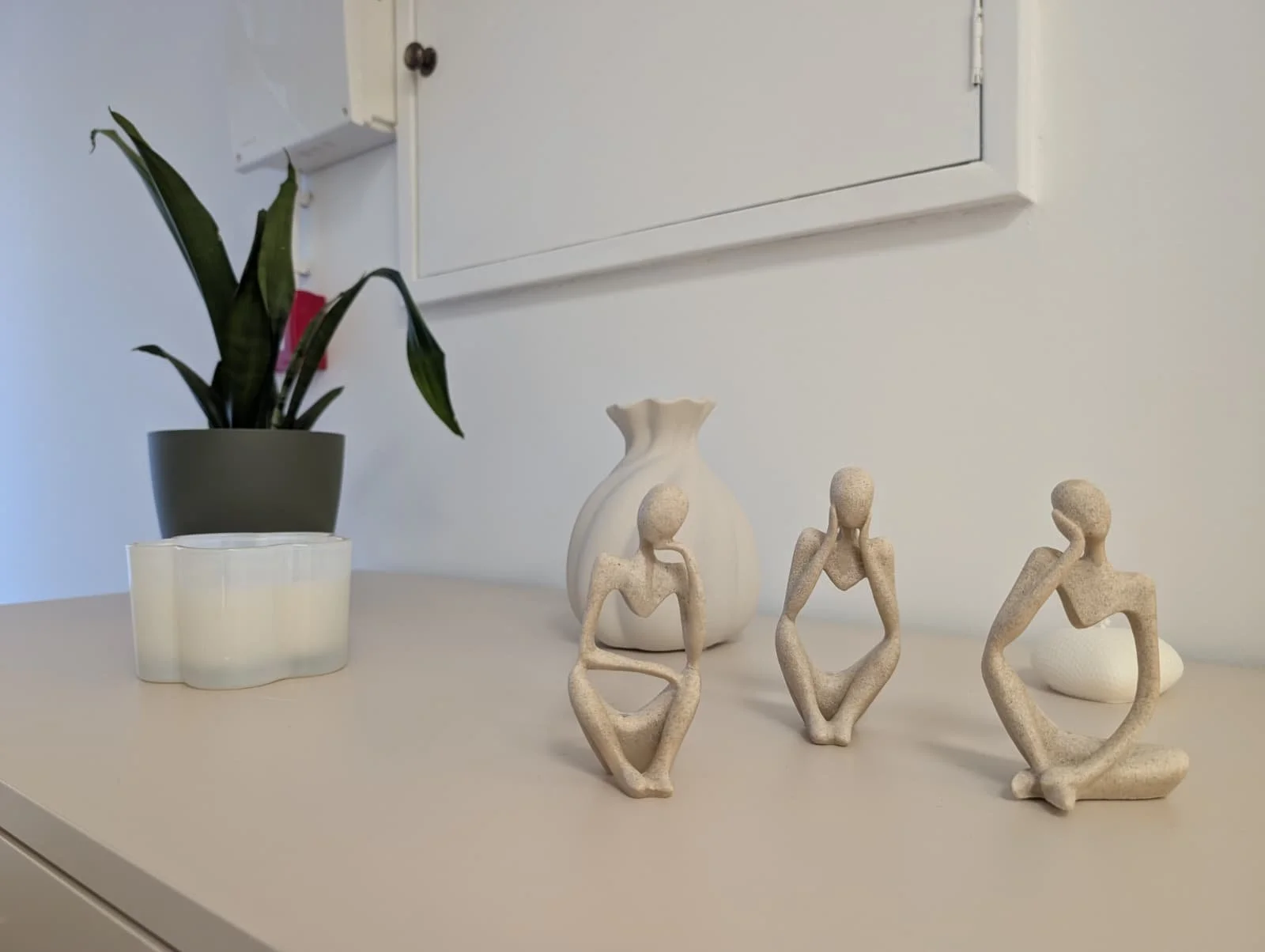 Decoração da Clínica Alma de mesa com uma planta em vaso, três estátuas de figuras humanas em posturas de reflexão ou meditação, uma vela decorativa e uma urna branca, tudo em ambiente com paredes brancas.