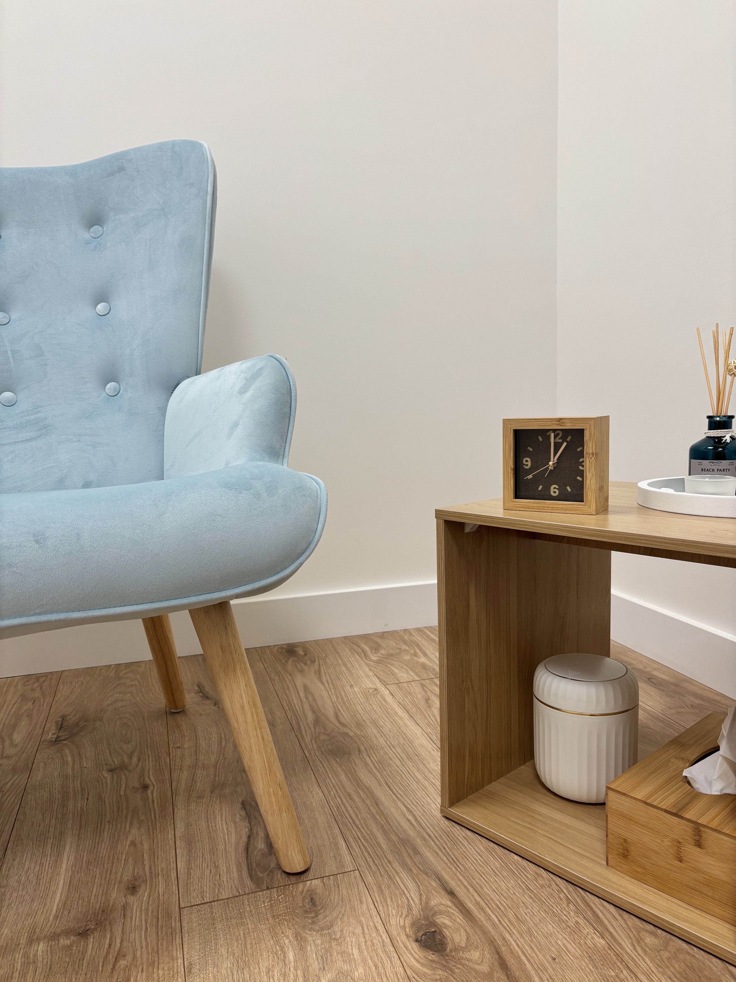 Consultório da Clínica Alma com uma poltrona azul com detalhes em madeira, uma mesa de canto de madeira com um relógio de mesa, e uma caixa com lenços de papel.