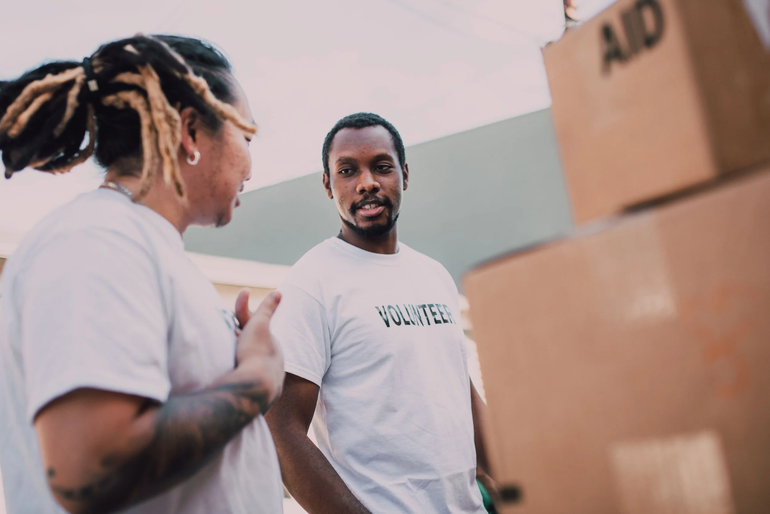 Two volunteers in white t-shirts talking beside stacked aid boxes at a community outreach event