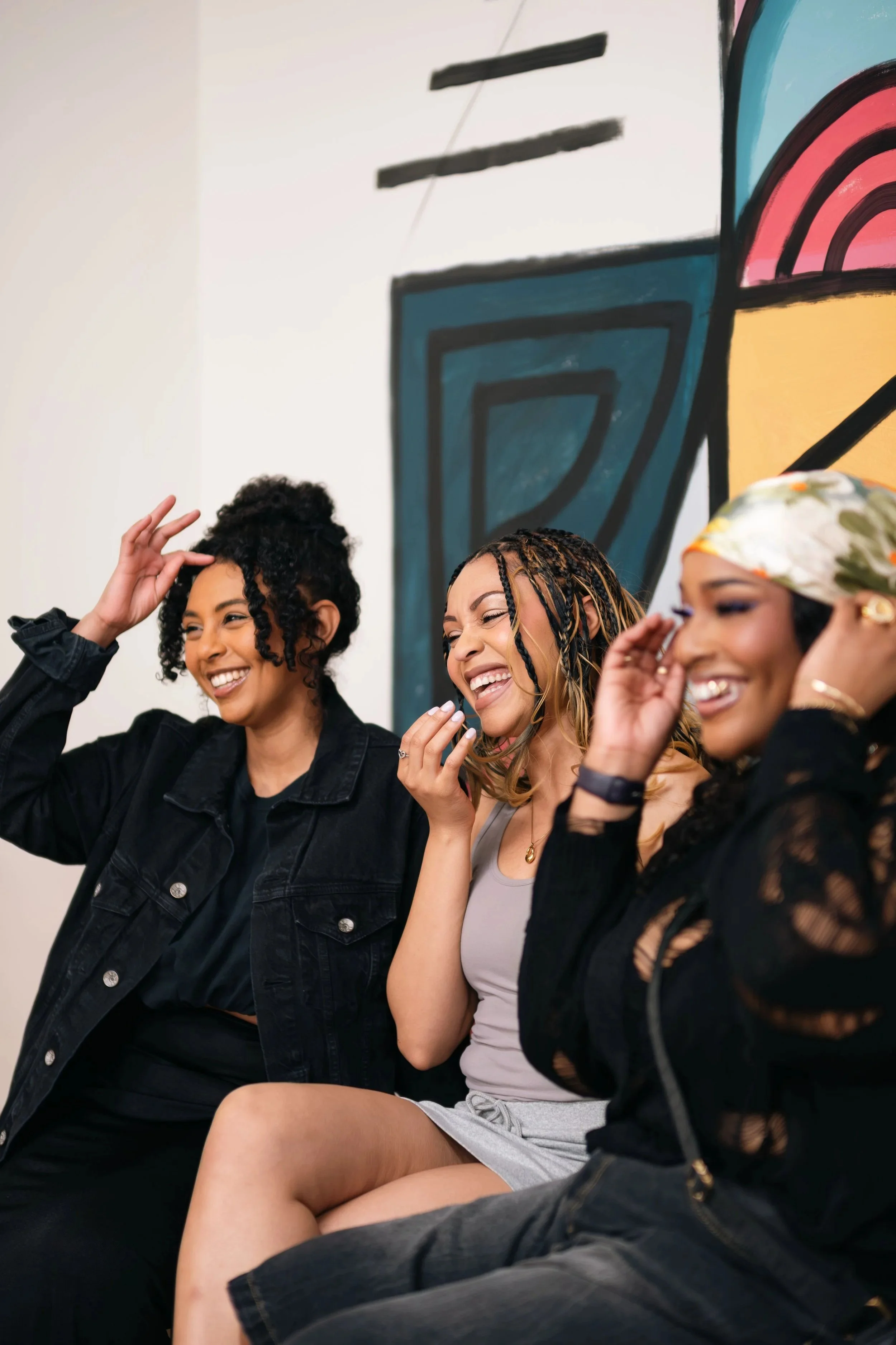 Three Black women laughing together while seated in front of a colourful abstract mural
