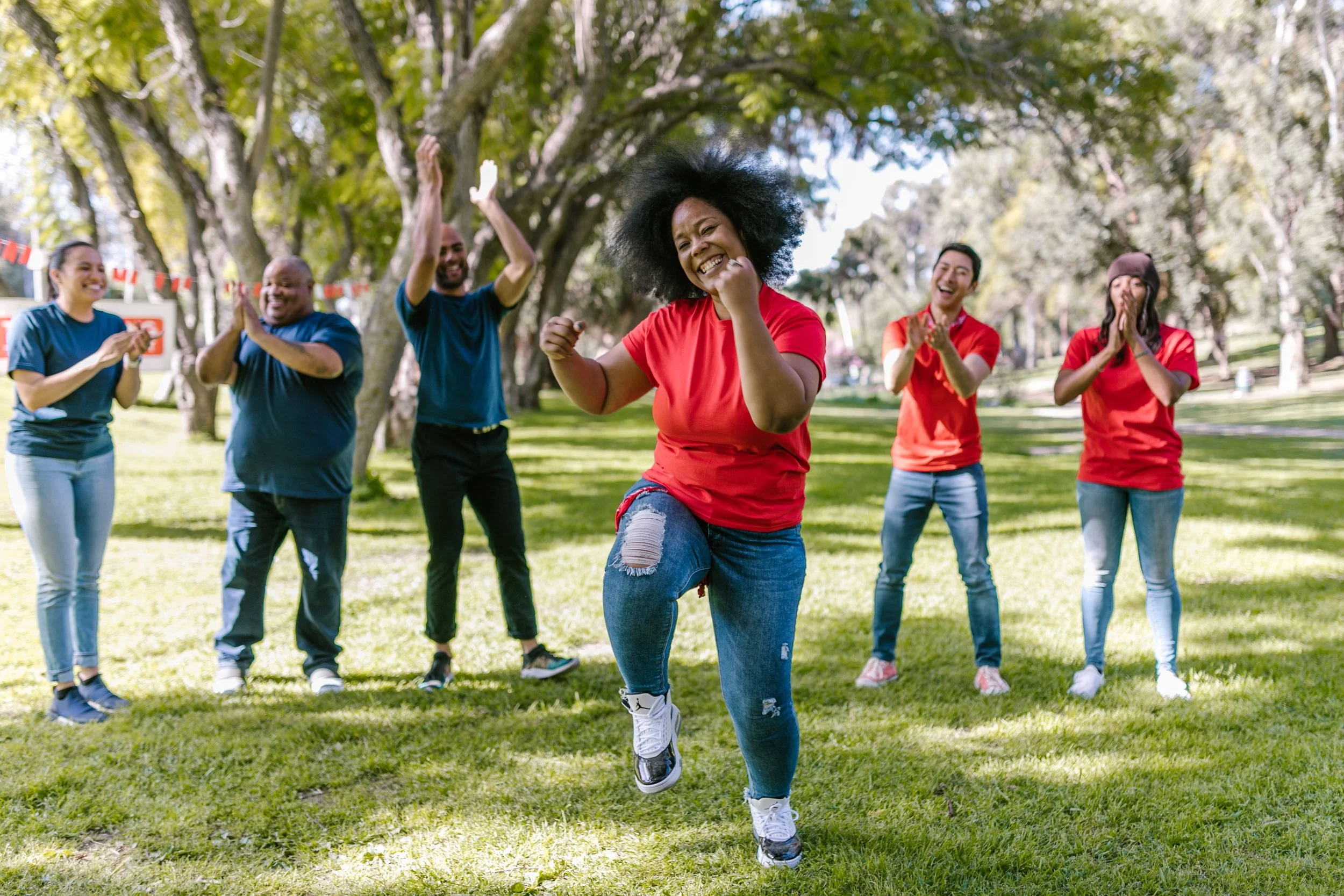 Woman dancing outdoors on grass while a diverse group cheers and claps around her