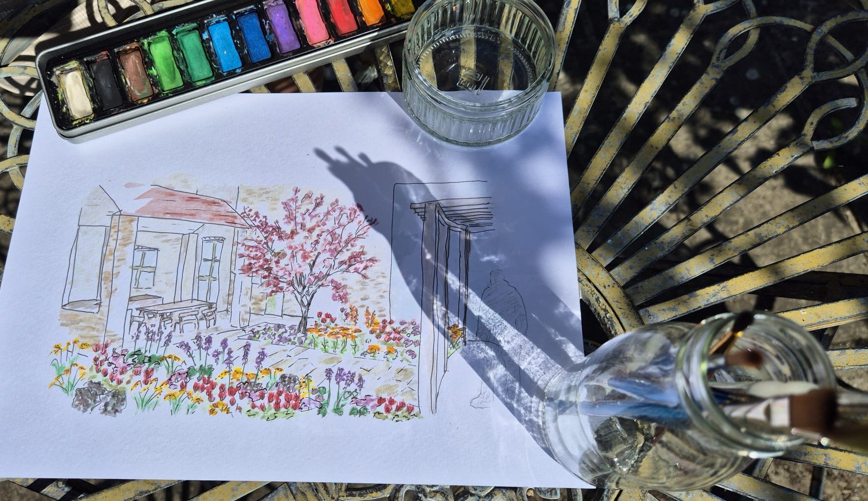 Watercolor sketch of a house with a garden of colorful flowers, a tree with pink blossoms, and outdoor furniture, placed on a white sheet of paper on a metal outdoor table. A glass of water, a jar with paintbrushes, and a watercolor paint set are also on the table.