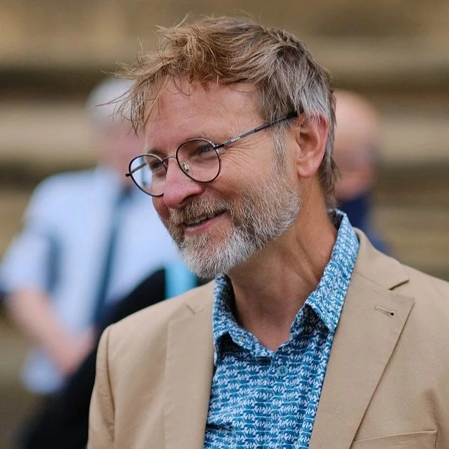 A smiling middle-aged man with glasses and a beard, wearing a beige blazer and a blue patterned shirt, outdoors at an event or gathering.