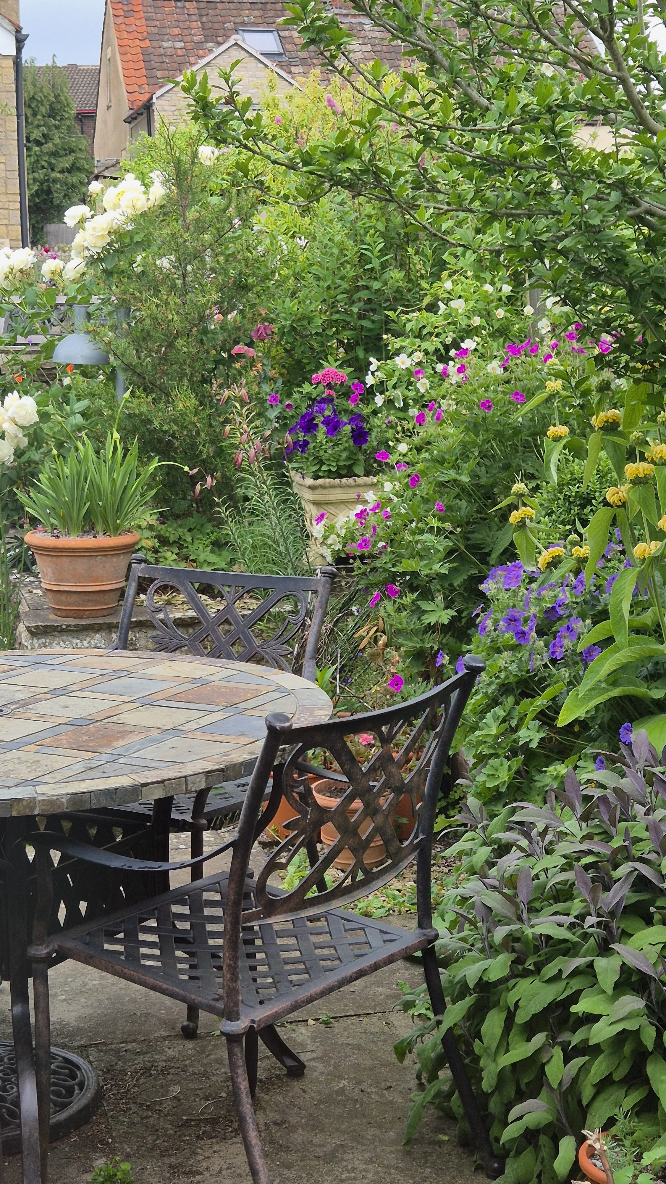 A garden with a round stone-tiled table surrounded by black wrought iron chairs, greenery and colorful flowers in pots and in the garden beds, with brick houses in the background.