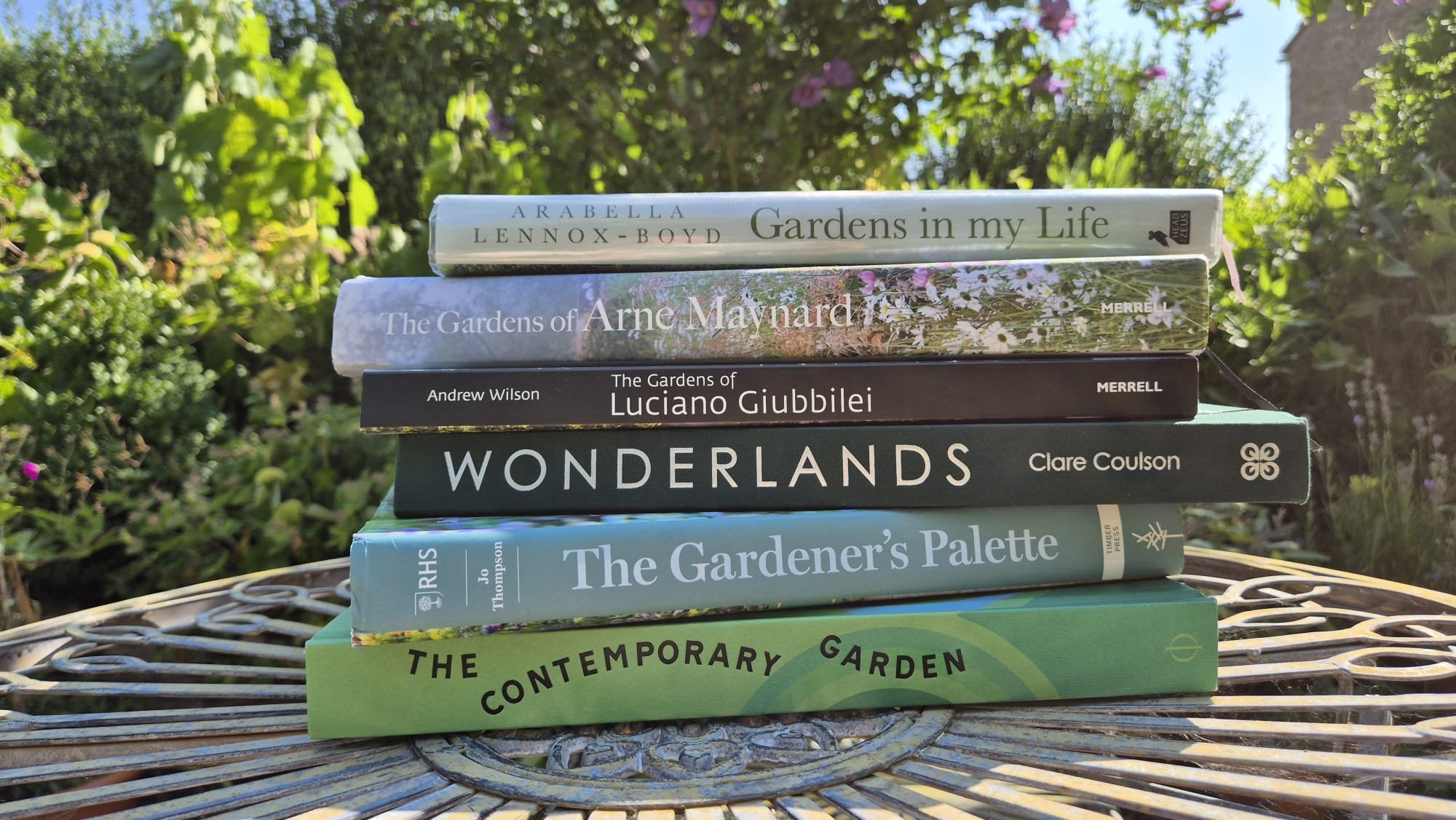Stack of seven gardening books outdoors on a wicker table with greenery and flowers in the background.