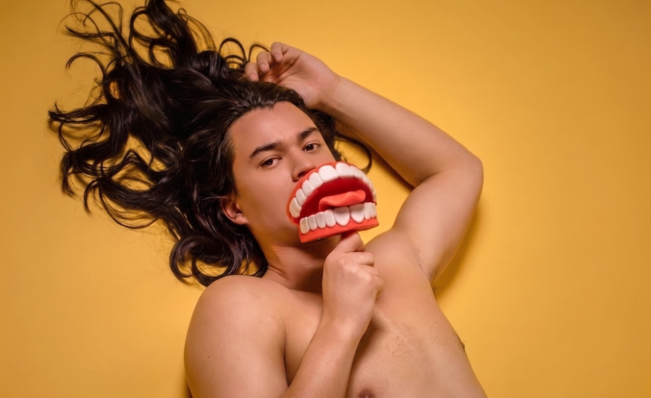 A person with long, curly dark hair lying on a yellow background, holding a large toy mouth with teeth and a tongue in front of their face, giving a playful expression.