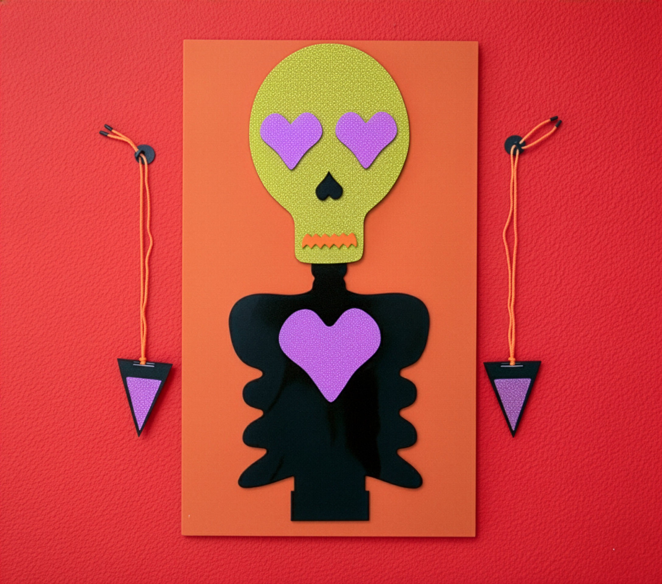 An art piece depicting a stylized skull with a green glittery surface, purple heart-shaped eyes, and an orange zigzag mouth, mounted on an orange background, with two small lanterns hanging on orange strings on either side.