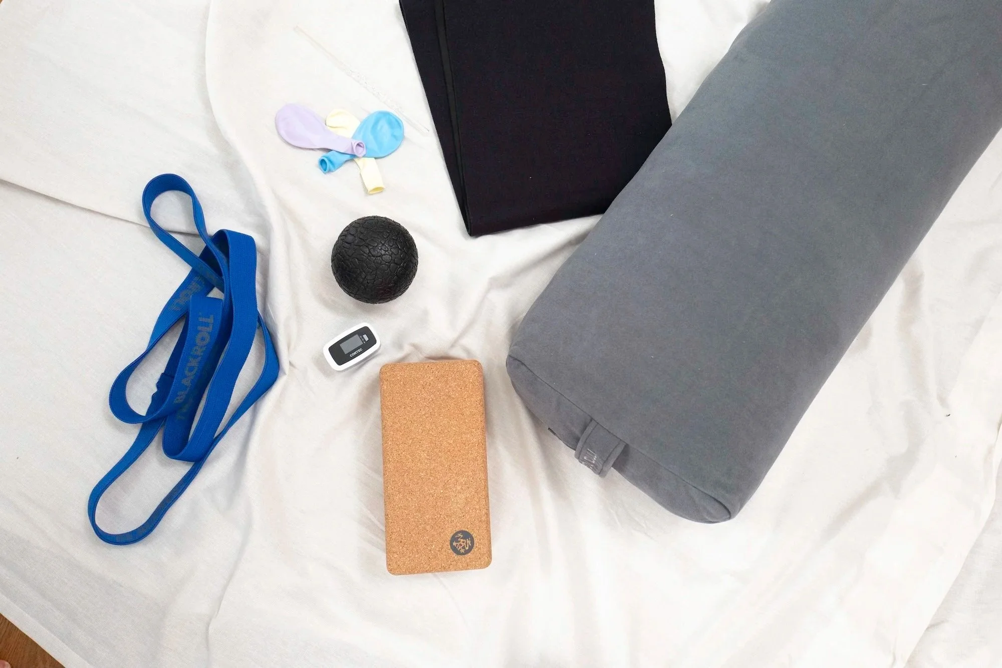 Verschiedene Gegenstände auf einem weißen Tuch, darunter ein blaues Widerstandsband, Pulsoximeter, Korkblock, schwarztexturierter Ball, grauer Gegenstand, schwarzer Waist-Trainer, bunte Ballons.