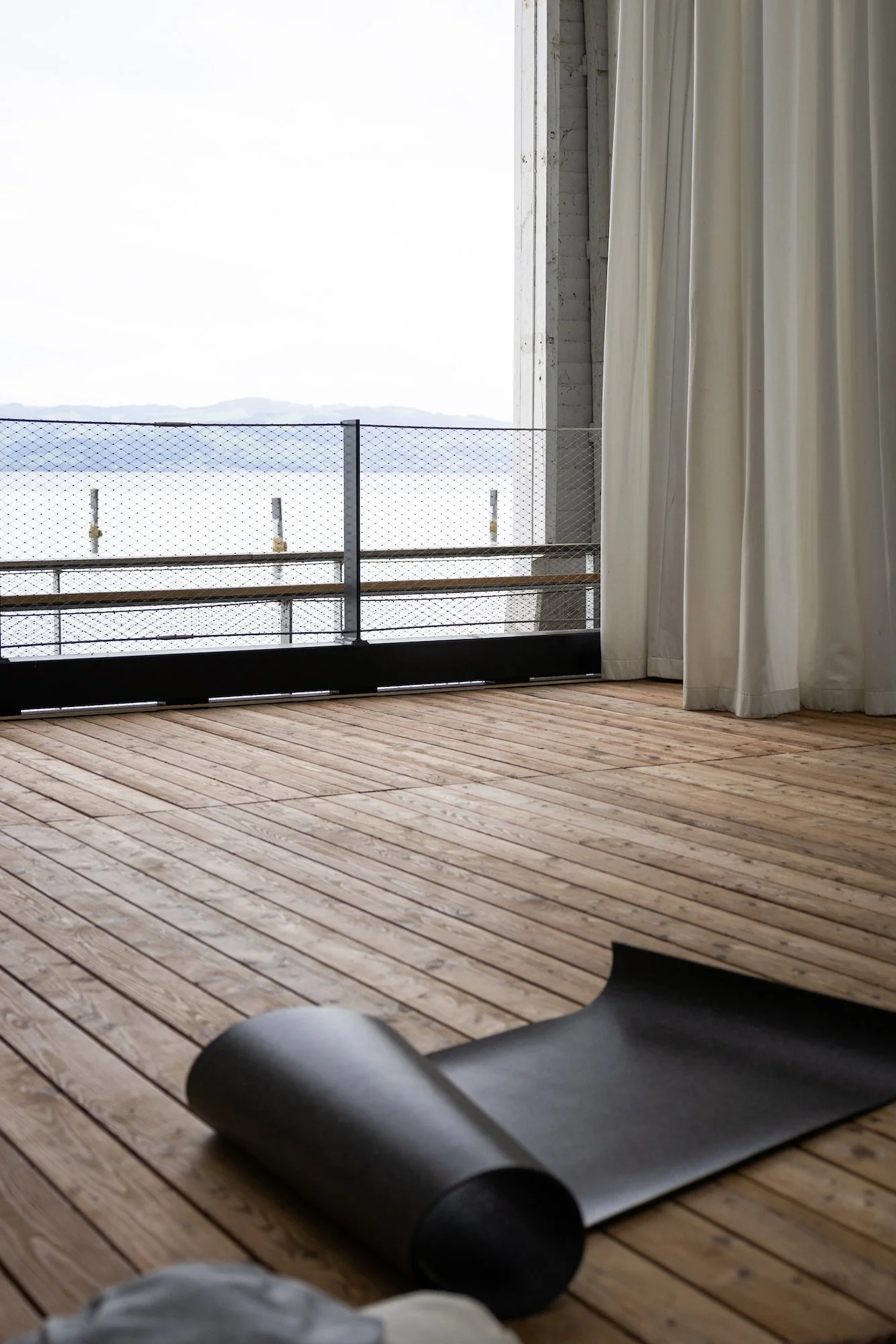 Ruhige Yogamatte auf einem Holzfußboden vor einem Balkon mit Blick auf See und Berge, teilweise von weißen Vorhängen verdeckt.