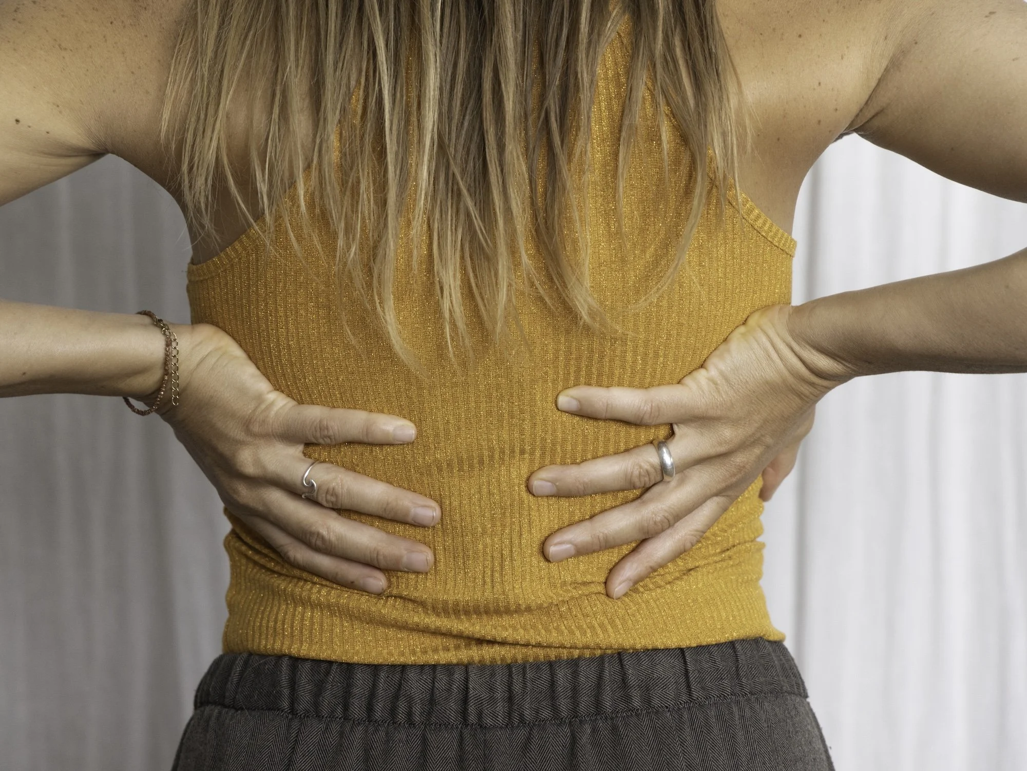 Nahaufnahme eines Frauen-Torsos, die Hände auf den Rücken gelegt, trägt ein orangefarbenes Top und braune Stoffhose.