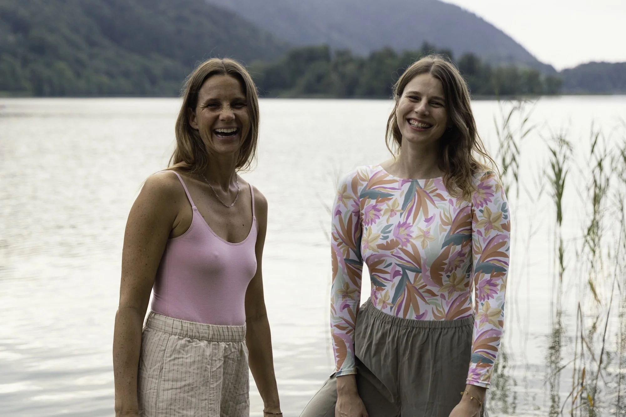 Zwei lachende Frauen stehen am Ufer eines Sees mit Hügeln im Hintergrund, während sie die Natur genießen.