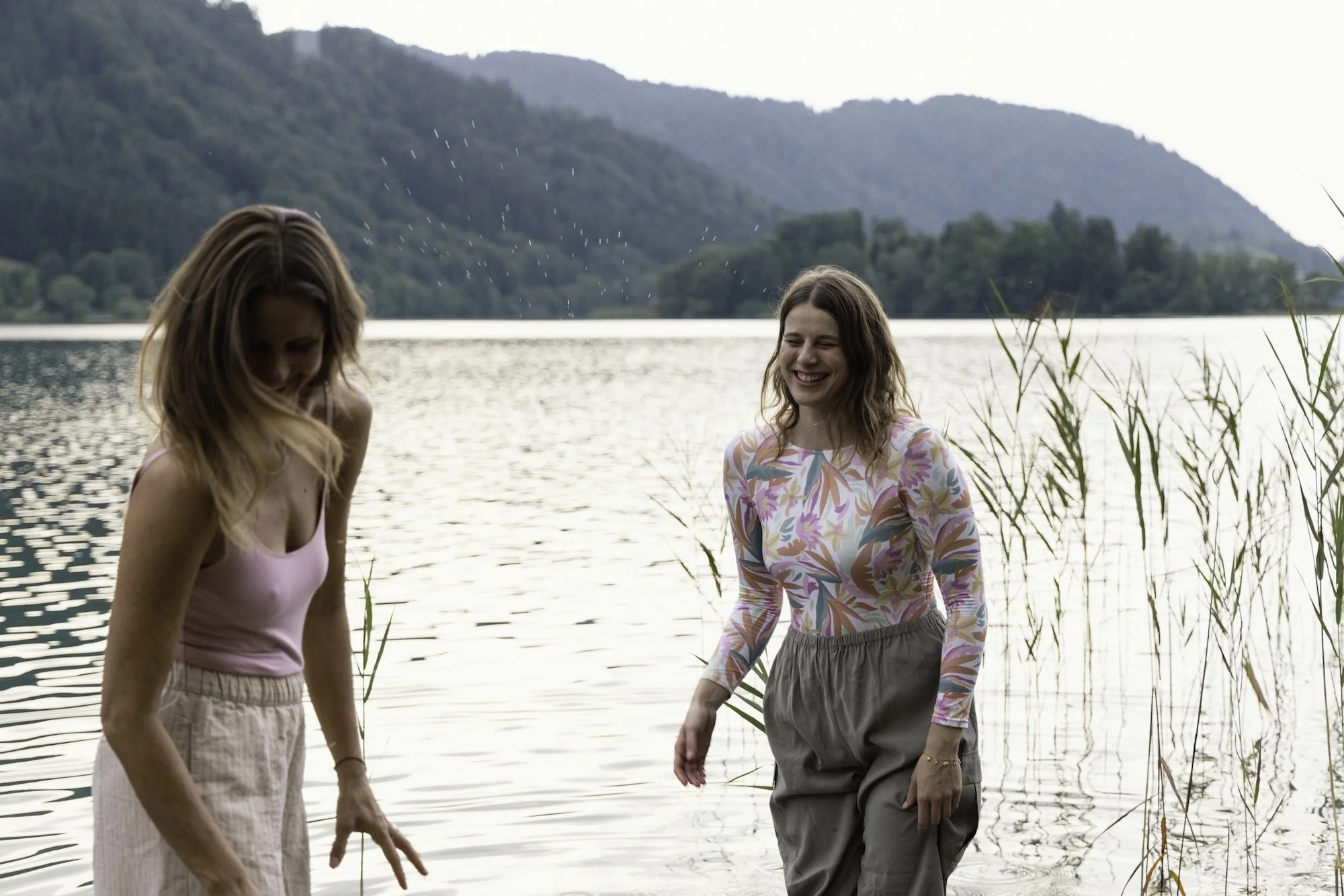 Zwei Frauen beim Spaziergang am See, lachend, umgeben von Natur, mit Wasser und Bergen im Hintergrund.