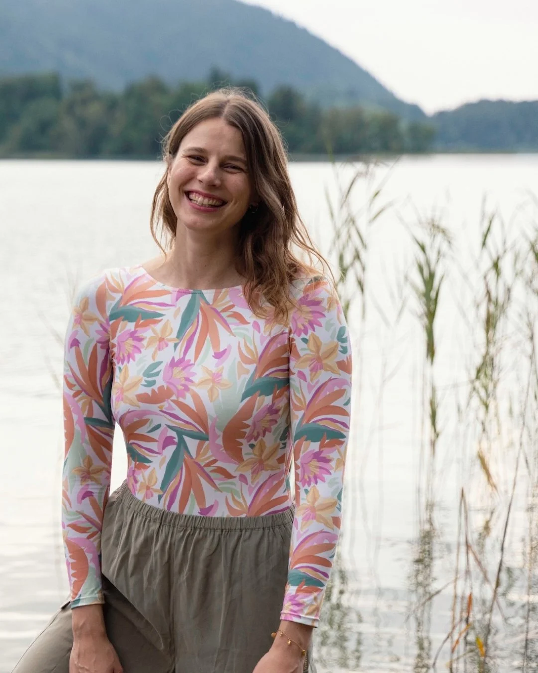 Junge Frau steht am Seeufer, lachend, mit Bäumen und Bergen im Hintergrund, trägt ein buntes Blümchenshirt und beige Hosen.