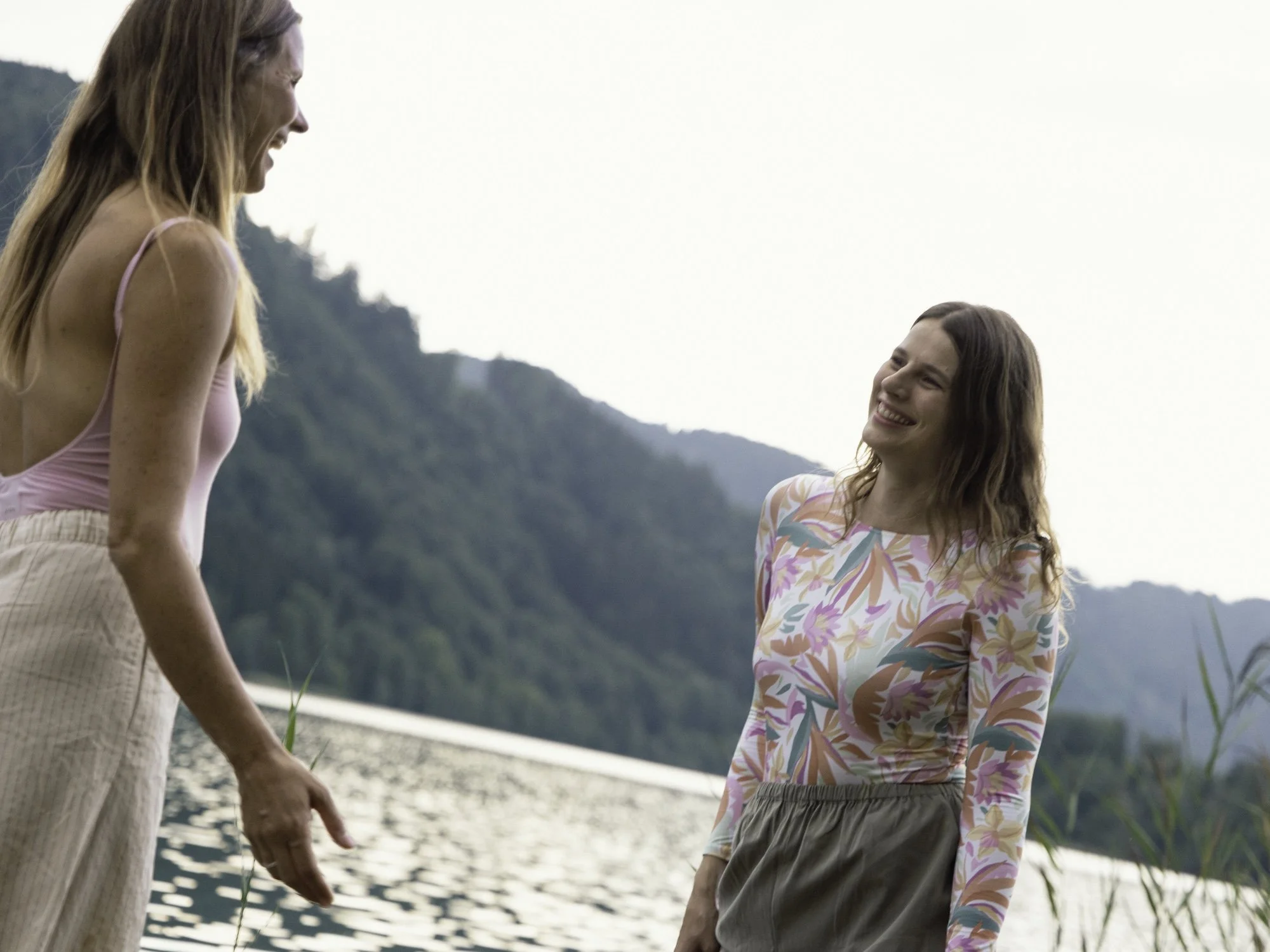 Zwei junge Frauen am Wasser mit Bergen im Hintergrund, lachen und haben Freude.