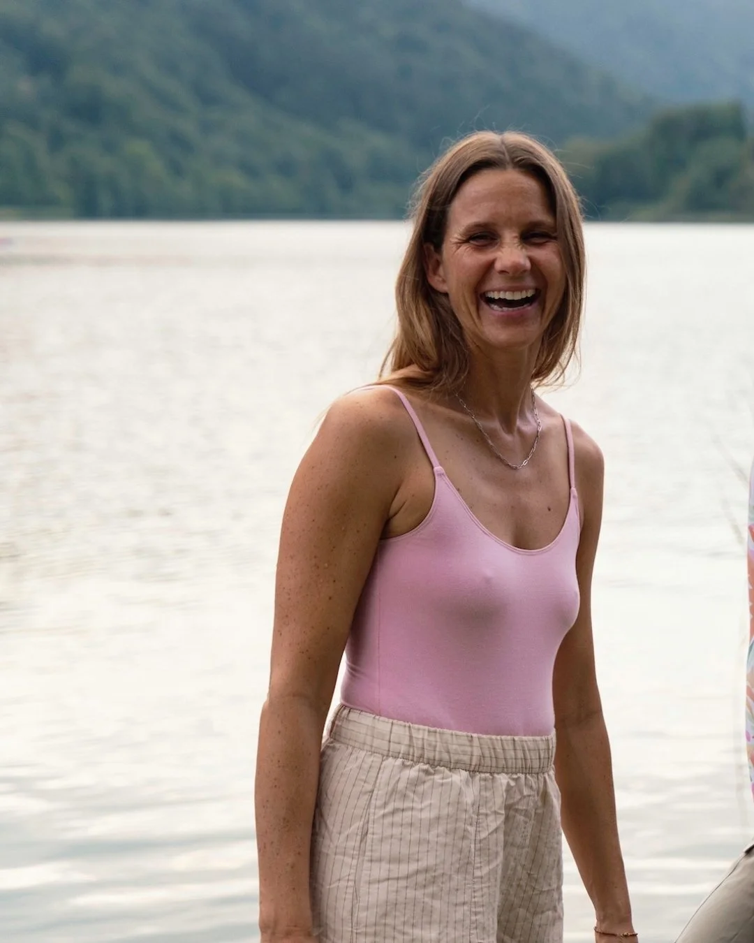 Eine lachende Frau mit braunen Haaren in einem pinken Top und hellen Shorts steht am Wasser mit Bergen im Hintergrund.
