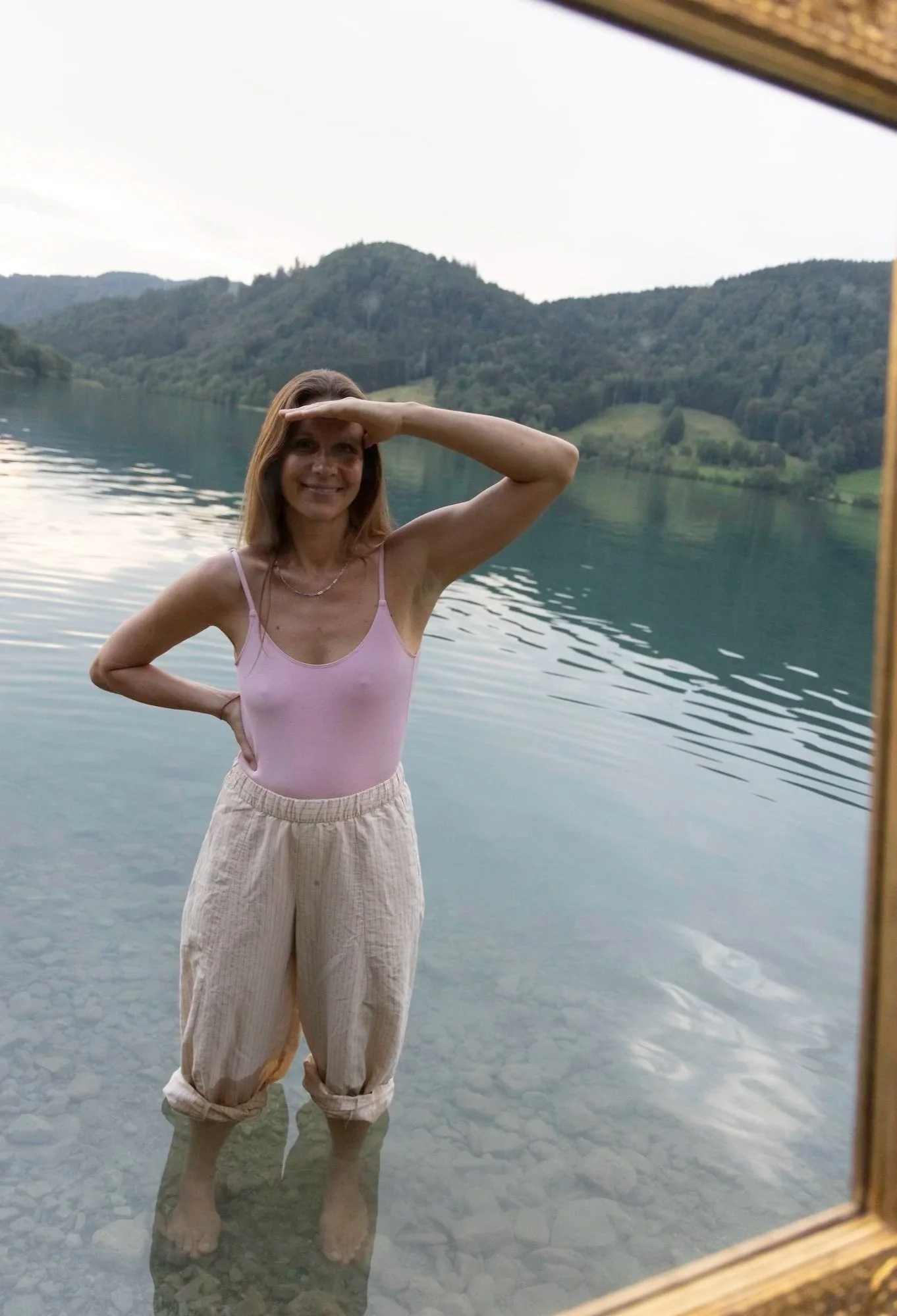 Eine Frau steht barfuß in einem klaren See mit einem bergigen, bewaldeten Hintergrund und blickt in die Ferne, während sie den Arm hoch hält, um die Sonne zu blockieren.