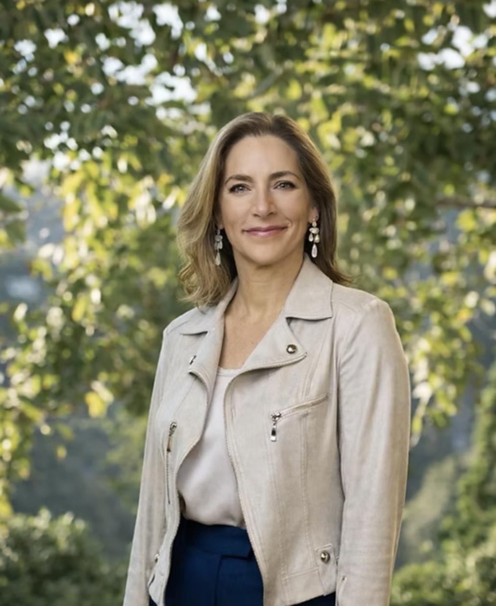 A photo of Rachel Goslins with shoulder-length brown hair wearing a beige leather jacket and white top standing outdoors with trees and foliage in the background.