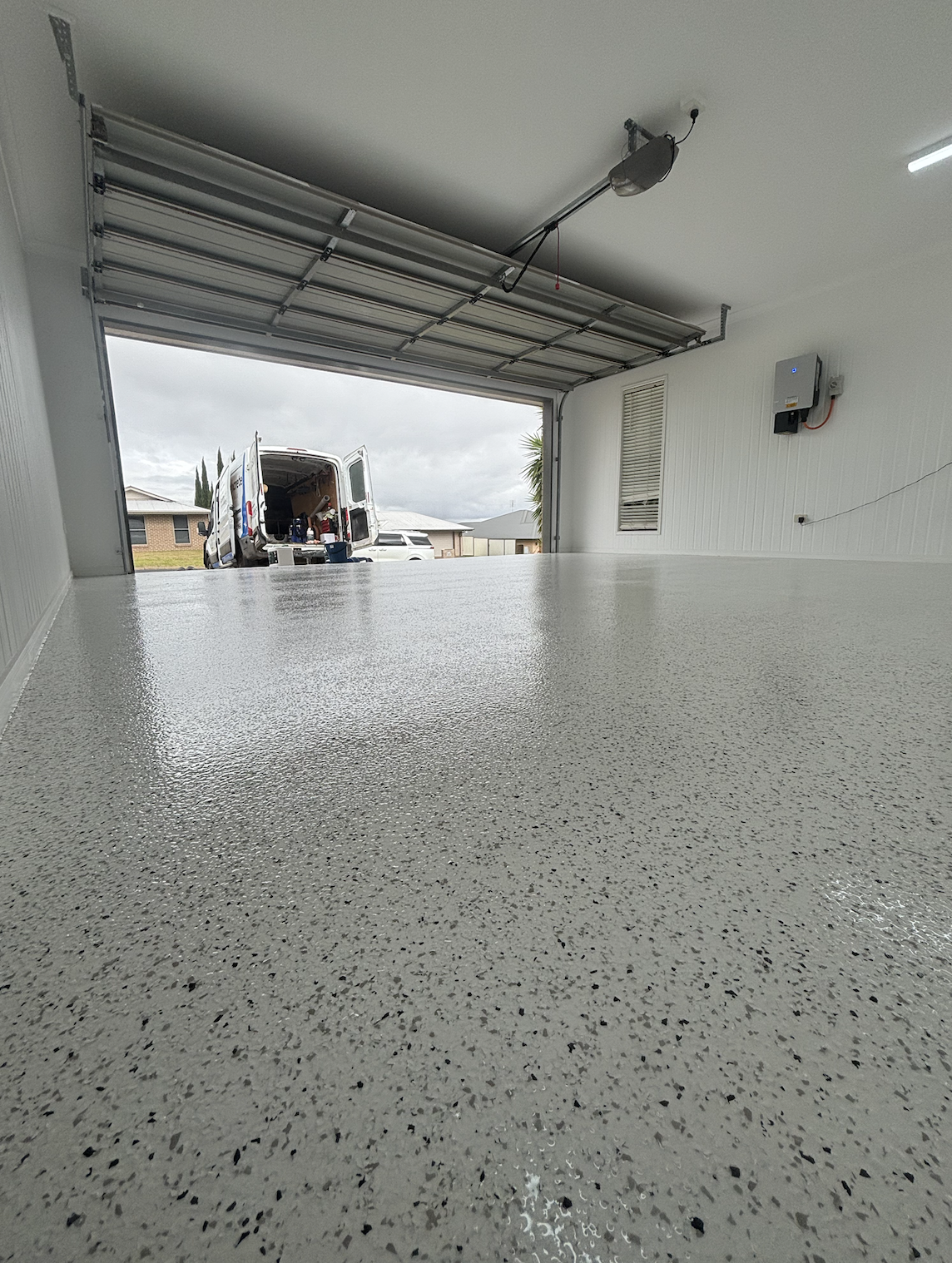View of an open garage with a newly epoxy-coated floor, looking out to a suburban street. A van is parked outside with its rear doors open.