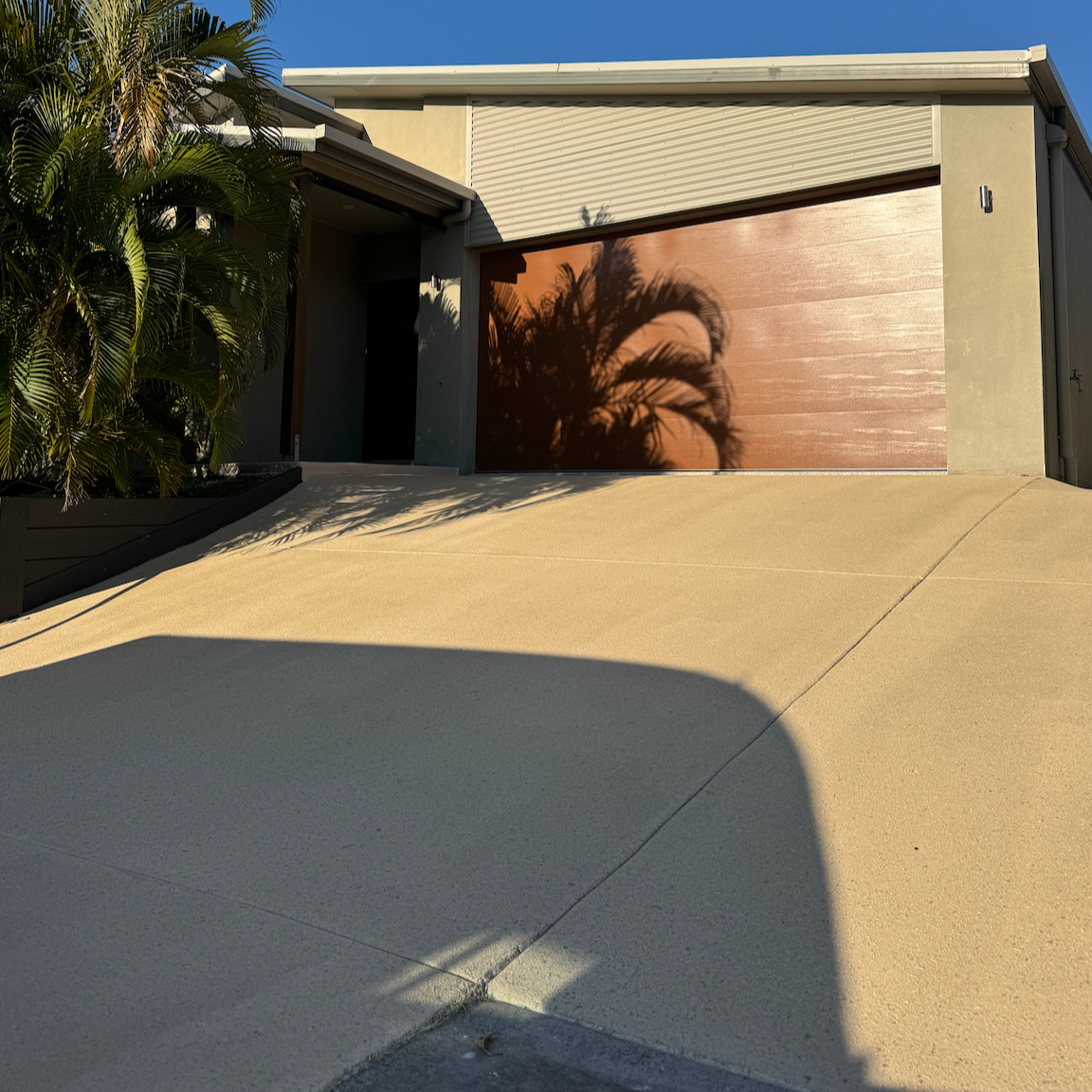 Exterior view of a modern house with a beige driveway, wooden garage door, and a palm tree casting a shadow on the door.