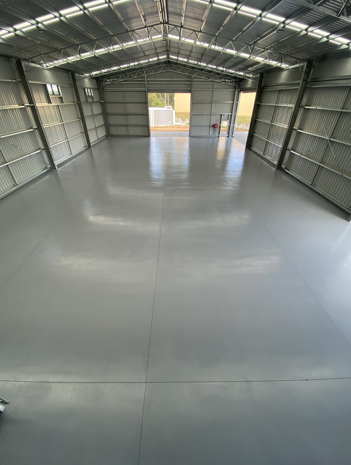 Empty metal warehouse with a concrete floor, overhead lighting, open garage doors, and outside view of trees and a building.