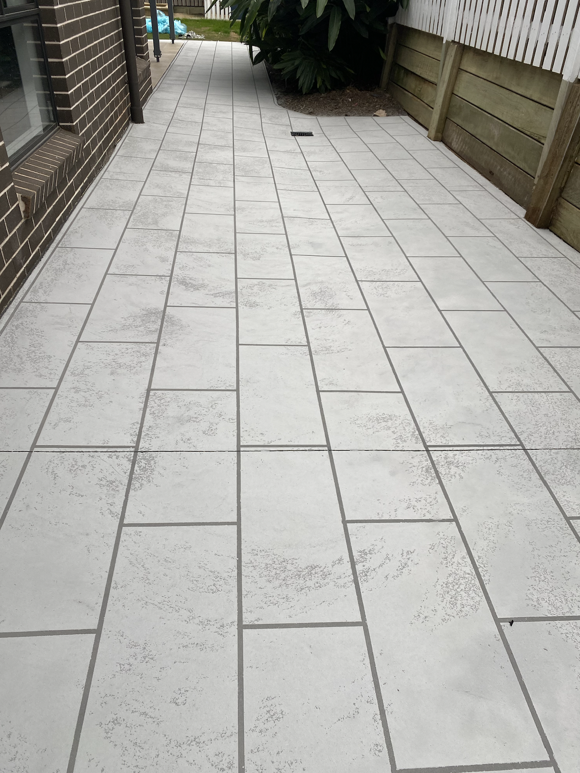 A tiled outdoor patio with light gray rectangular tiles, a drainage grate, green plants, a wooden fence, and a brick house wall.