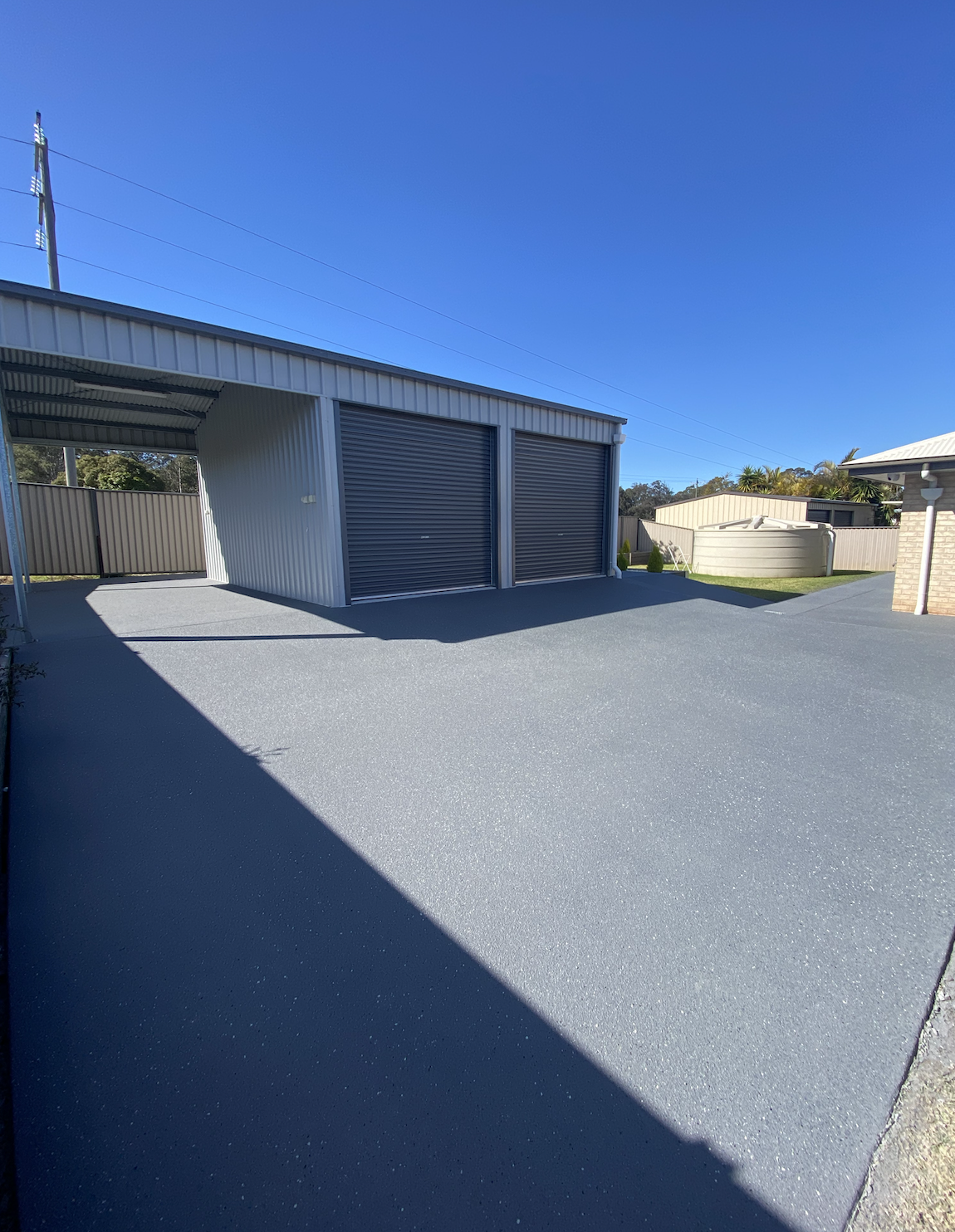 A modern backyard with a gray concrete patio, a metal shed with roller doors, a water tank, and a bamboo fence under a clear blue sky.