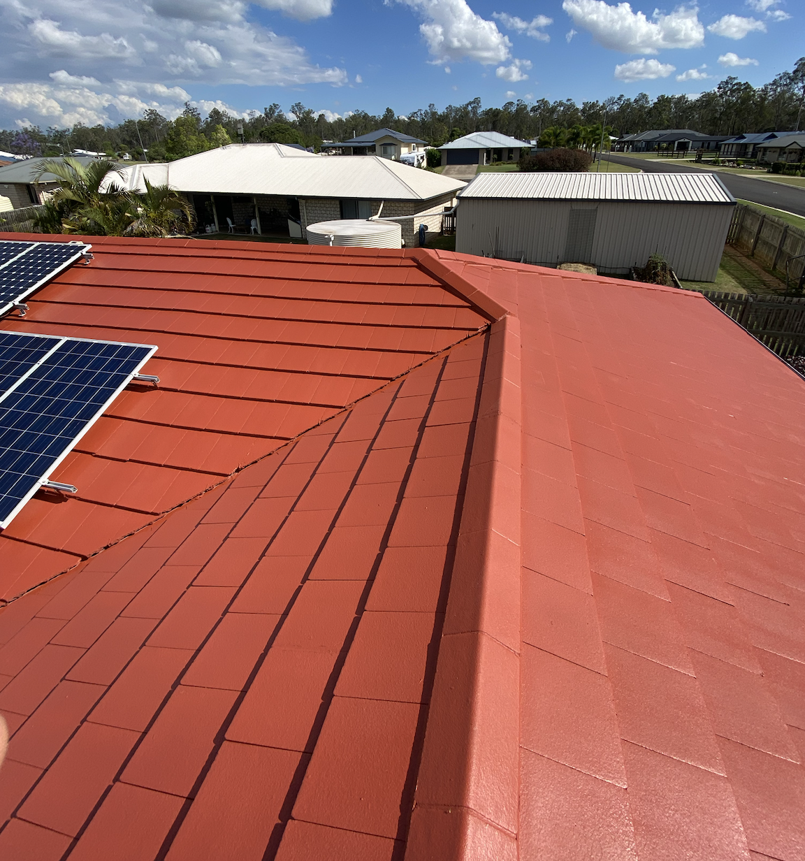Red tiled roof with solar panels on the left, in a neighborhood with nearby houses, a water tank, and trees in the background.