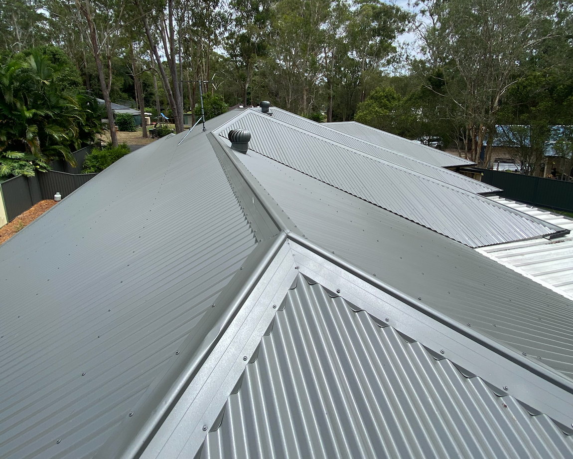Multiple metallic, corrugated roofing sheets installed on a building, with trees and a cloudy sky in the background.