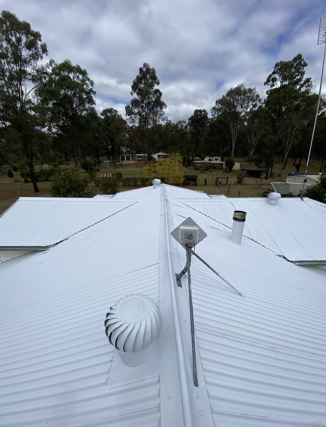 View from above of a white sheet metal roof with vents, a solar panel, and a chimney, with trees and houses in the background and a cloudy sky.