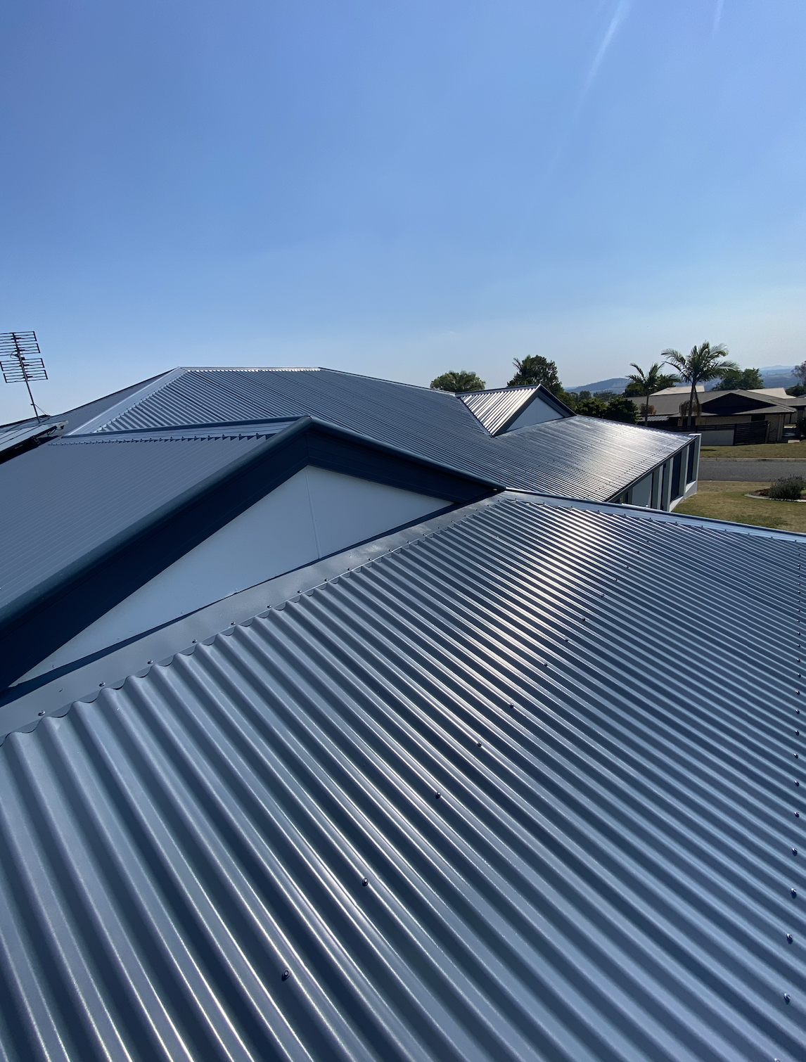 Close-up of silver corrugated metal roof reflecting sunlight, with neighboring rooftops and a clear blue sky in the background.