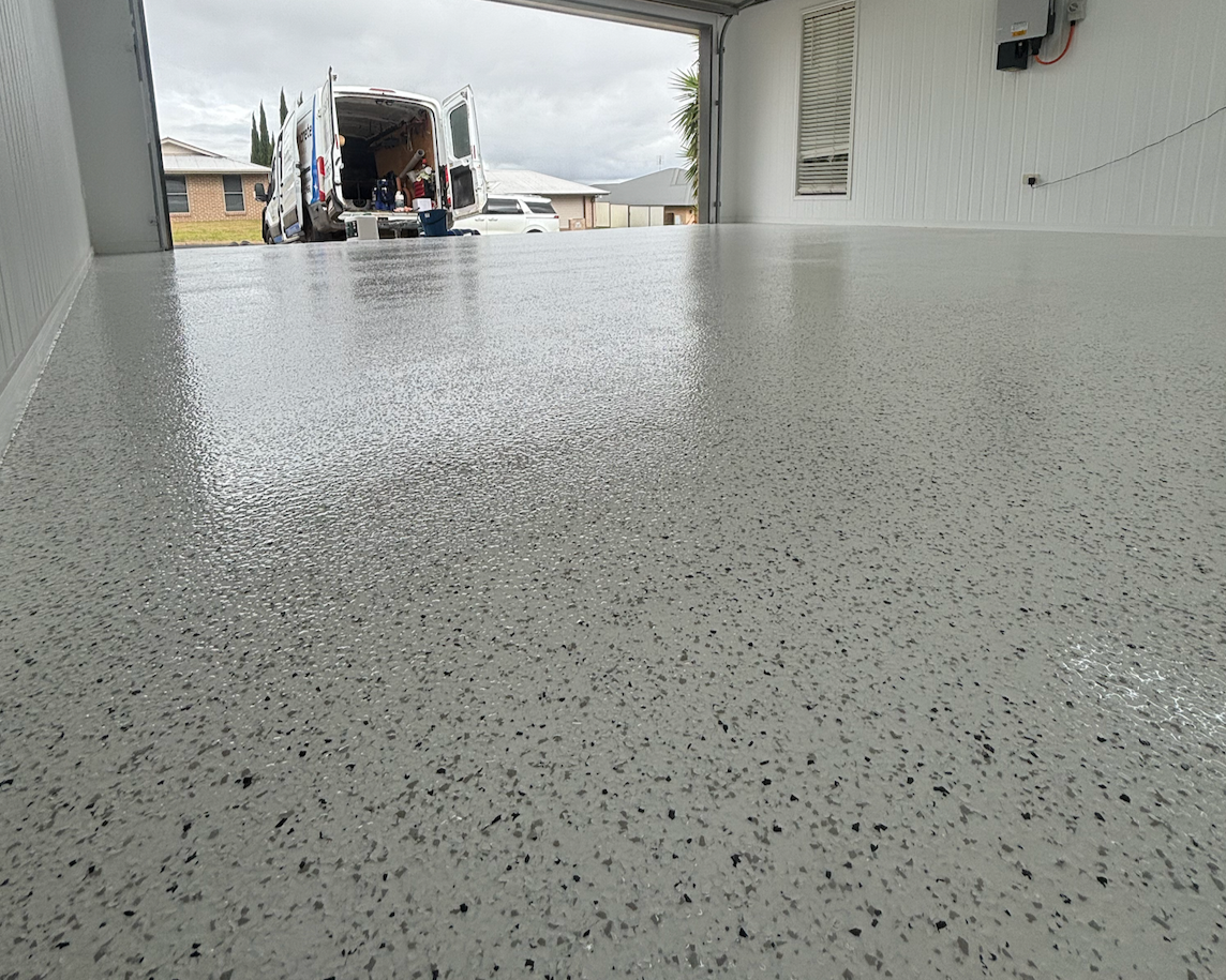 Freshly coated epoxy garage floor inside a residential garage with an open door, showing a van and a house outside on a cloudy day.