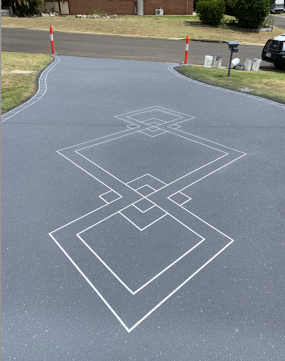 A small outdoor basketball court with white boundary lines and markings on a dark gray surface, bordered by grass and sidewalk. The court is partially blocked by orange traffic cones.