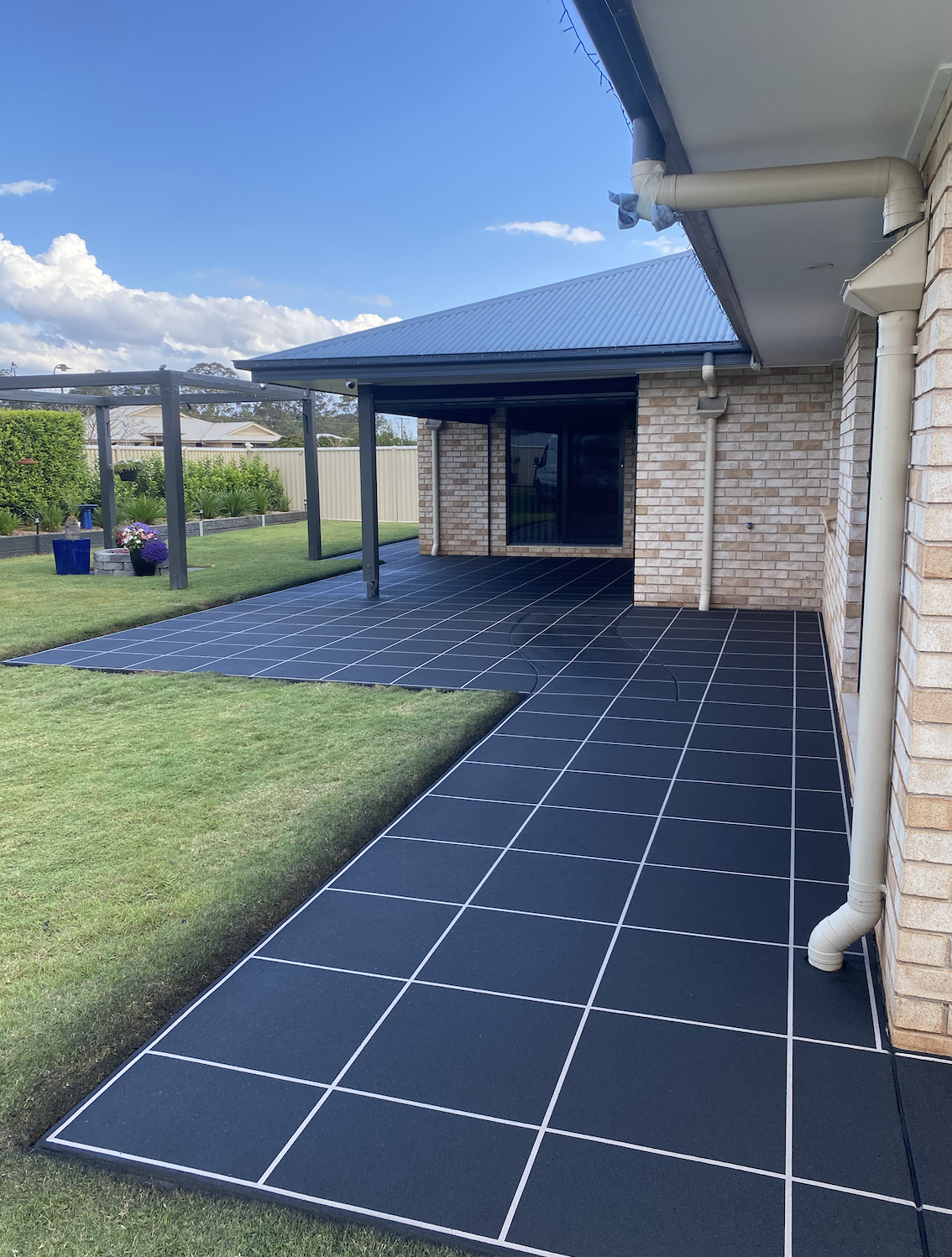 Backyard with tiled patio, lawn, and garden area, brick house with gutter system, blue sky with clouds.