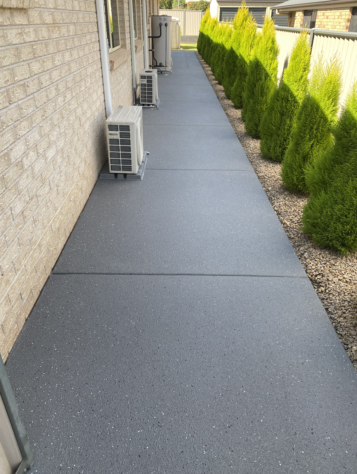A backyard patio with textured gray concrete slabs next to a brick house. Green shrubs line the right side, separated from the patio with small rocks. Air conditioning units are positioned against the house wall.