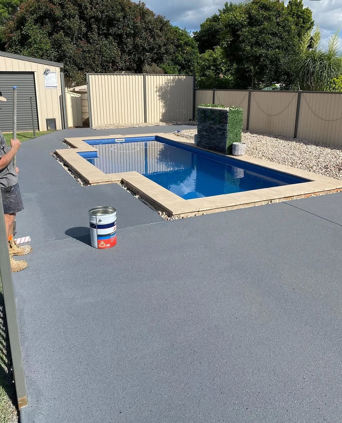 A backyard with a rectangular swimming pool, surrounded by a concrete deck, next to a privacy fence and trees; a water feature with a stone wall is at one end of the pool.