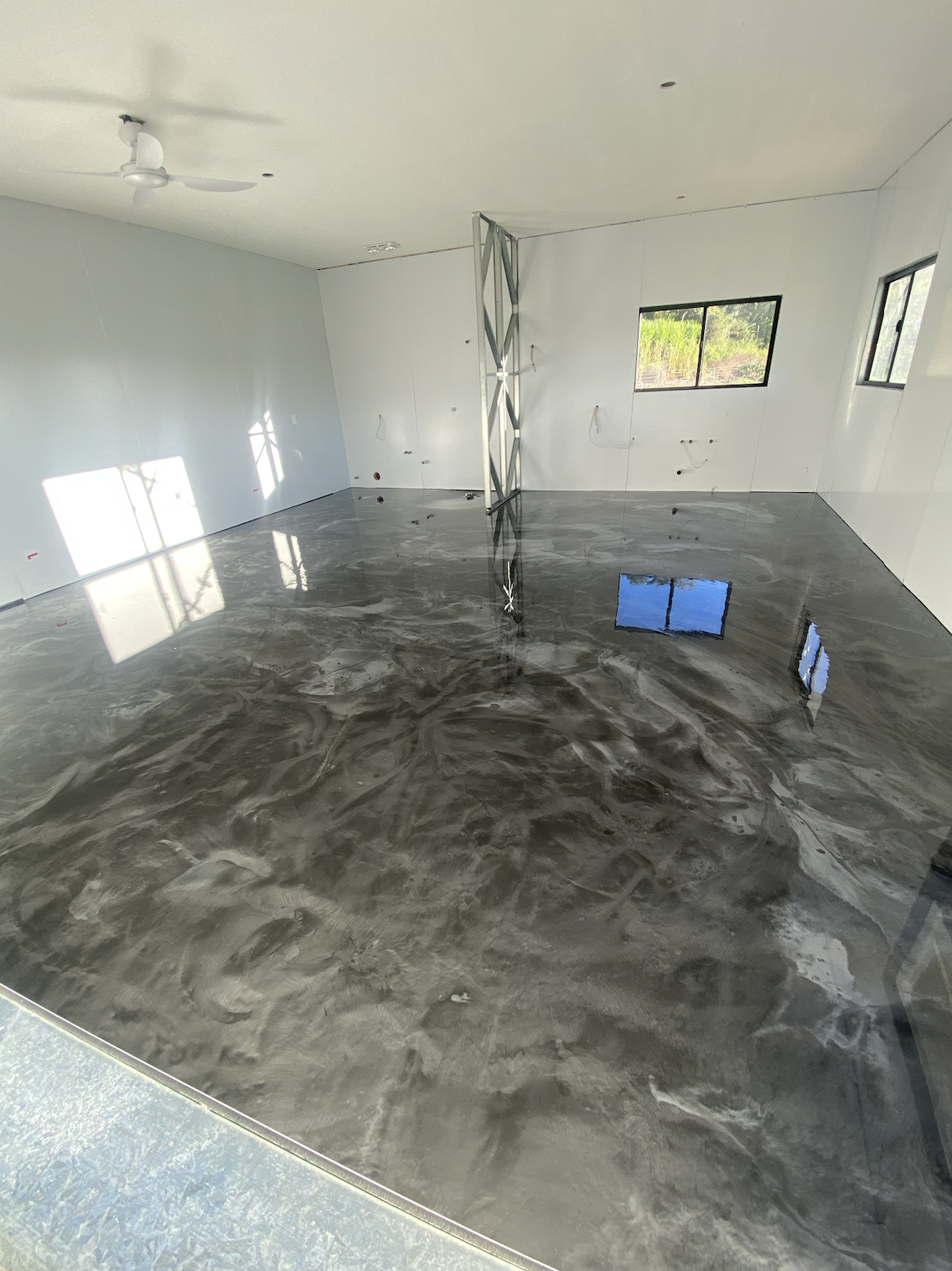 Interior of a new construction room with freshly poured concrete floor, white walls, and two windows, with a ceiling fan and construction support beams.