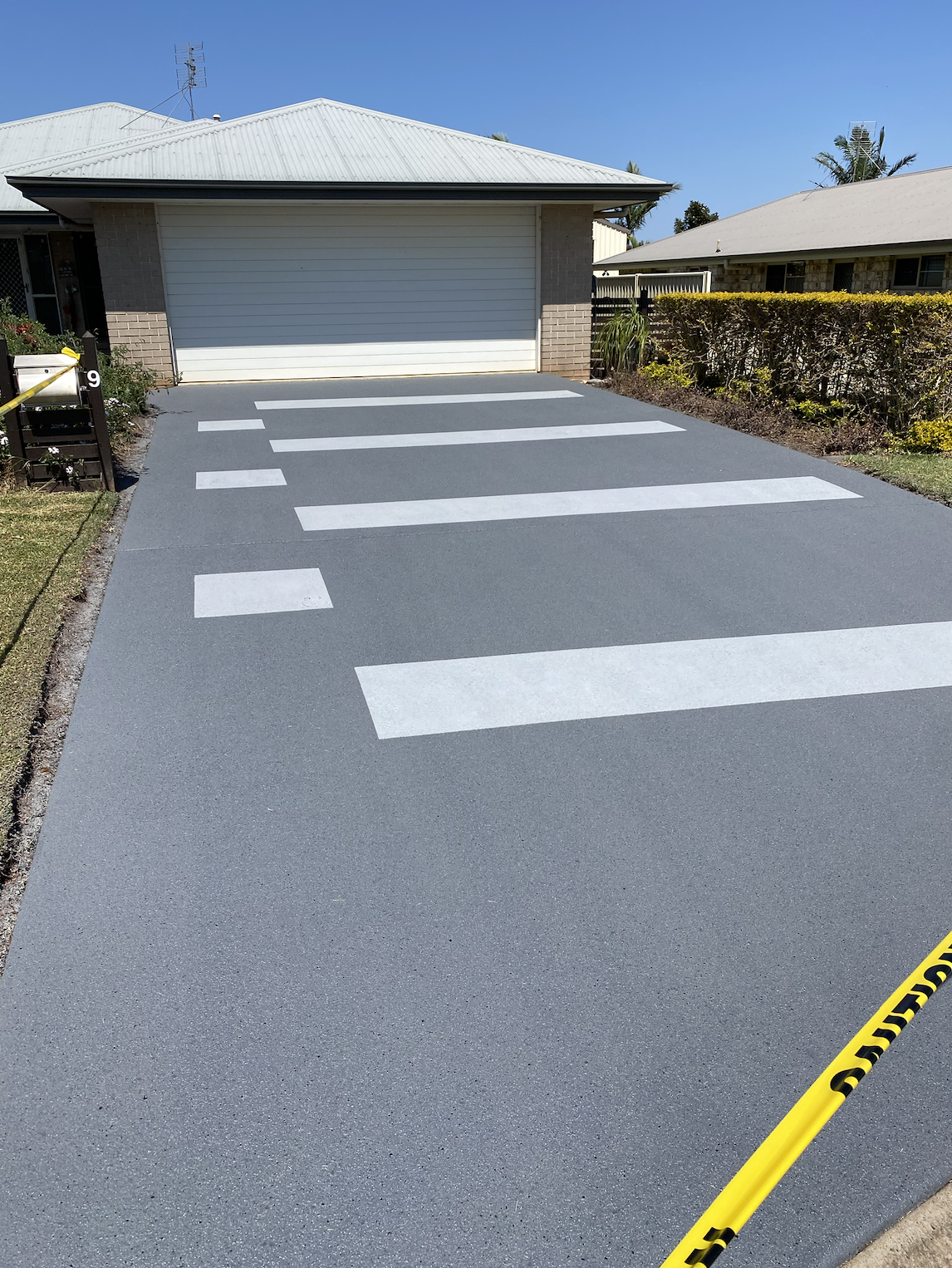 A newly painted driveway with white crosswalk lines leading up to a garage. Caution tape is visible on the bottom right corner, blocking part of the driveway.
