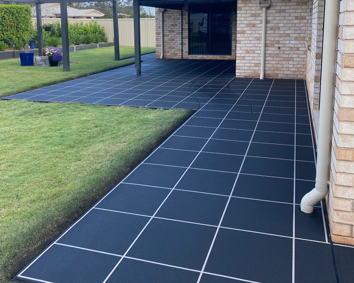 Black tiled patio with white grout, adjacent to a brick house with a gutter pipe, and a grassy yard with flowers and plants in pots.
