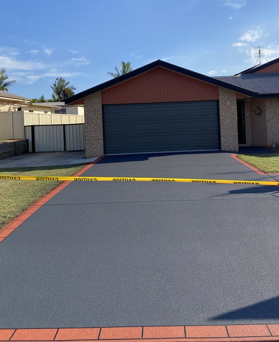 A house with a blue garage door and a driveway marked with yellow caution tape. The driveway is bordered with red bricks and the house has a brick exterior with a tiled roof. The sky is clear blue with some clouds, and there are palm trees in the bac