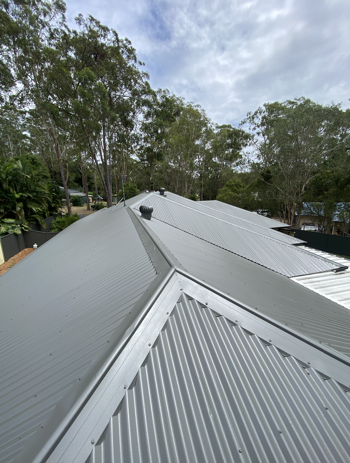 Multiple metal roofs with ridges and vents, surrounded by trees with a partly cloudy sky overhead.