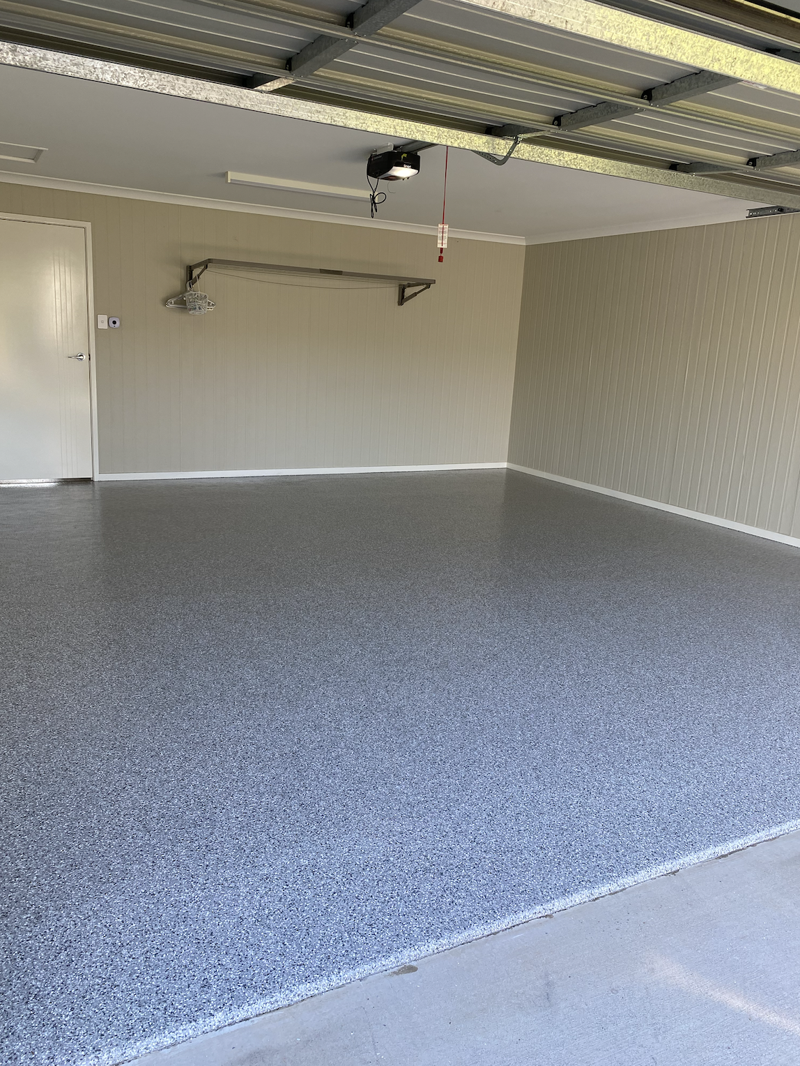 An empty garage with a textured grey floor, beige paneled walls, a closed white door, a wall-mounted metal rack, and a garage door opener mounted on the ceiling.