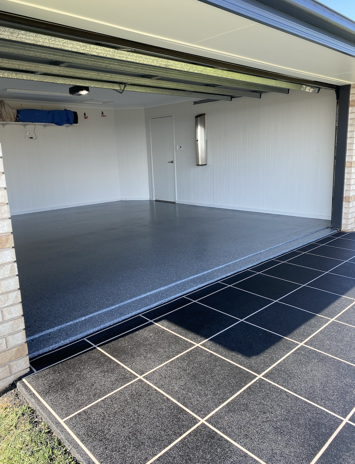 Open garage with a black tiled driveway outside, showing a gray inner floor, white wall panels, a closed door, and two small red hooks on the wall.