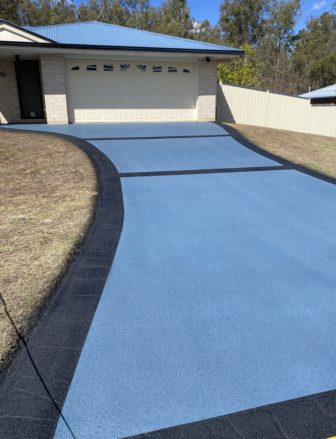 A blue driveway leading to a white garage with windows in the door, surrounded by a yard with brown grass and a beige fence, with trees and a blue sky in the background.